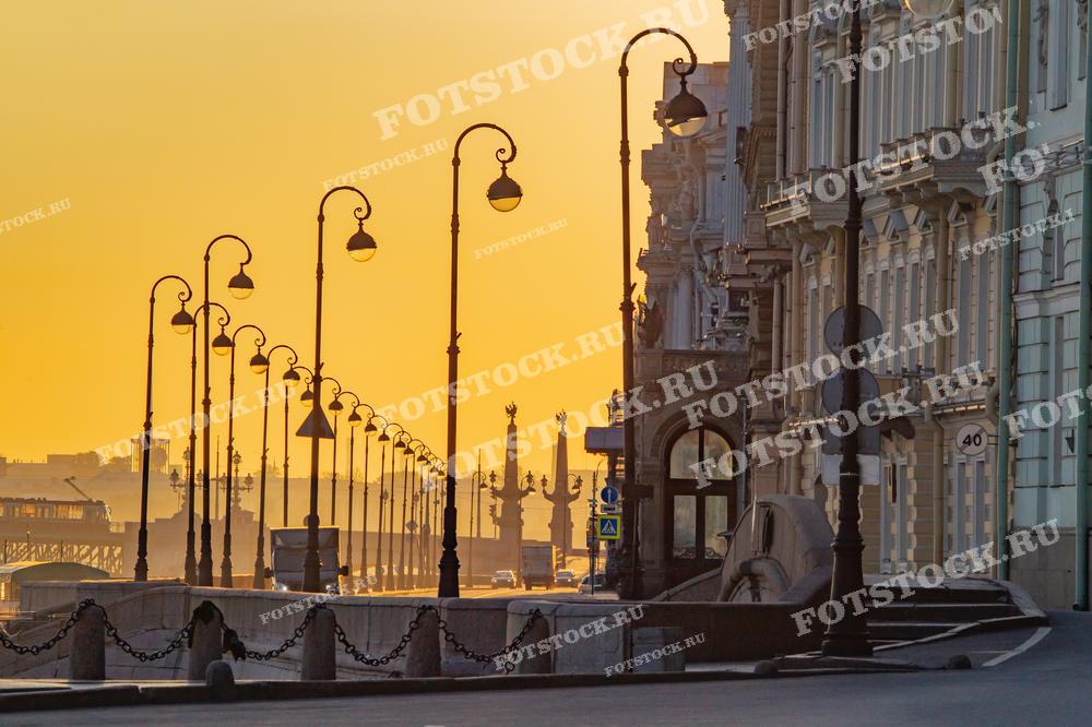 На фотографии изображена набережная Санкт-Петербурга ранним утром. Старинные фонари выстраиваются в ряд вдоль улицы, создавая эффект перспективы. Рассветное освещение окрашивает город в теплые оттенки желтого и оранжевого, подчеркивая красоту архитектуры. Тишина и спокойствие утреннего города передают атмосферу умиротворения.
